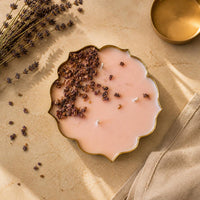 A scalloped gold metal candle dish filled with pale pink wax, topped with dried flower buds, styled on a beige textured surface with lavender sprigs and a small gold bowl nearby.