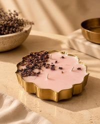 A scalloped gold metal candle holder filled with pale pink wax, topped with dried purple flower buds, styled on a beige stone surface with warm natural light and soft-focus decorative props.