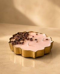 A scalloped gold metal candle holder filled with pale pink wax, topped with dried flower petals and two white wicks, set on a warm beige surface with soft sunlight and shadows.