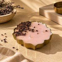 A scalloped gold metal candle holder filled with pale pink wax, topped with dried lavender buds, styled on a beige fabric surface with warm sunlight and neutral props.