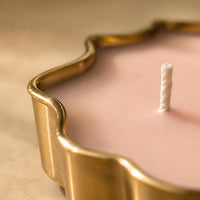 Close-up of a pink wax candle in a scalloped gold metal container with a single white wick, set against a warm beige background.
