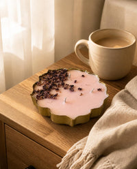A scalloped gold candle tin filled with pale pink wax and topped with dried purple flower petals, styled on a wooden bedside table next to a ceramic mug and a cream throw blanket.