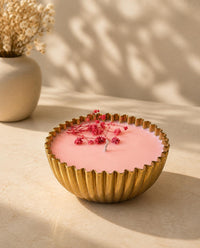 A pink wax candle in a fluted gold metal bowl, decorated with small red berries, set on a beige surface with warm sunlight and shadows.