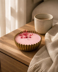 A pink wax candle in a fluted gold metal cup sits on a wooden bedside table next to a ceramic mug and a beige linen throw, with soft sunlight and window curtains in the background.