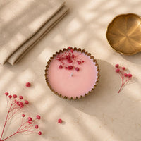 A pink scented candle in a scalloped brass vessel, styled on a beige marble surface with dried pink flowers, a folded beige linen cloth, and a gold-toned dish.