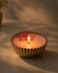 A lit candle in a ribbed metallic gold bowl with small red berries on the surface, set on a warm beige surface with soft shadows.