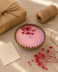 A pink wax candle in a scalloped gold metal cup with dried red flowers on top, styled on a linen surface with a kraft gift box, twine, a blank card, and scattered red flowers.