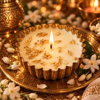 A lit cream-colored candle in a scalloped gold metal holder, decorated with dried flower petals, set on an ornate gold tray with white flowers and warm bokeh lights in the background.