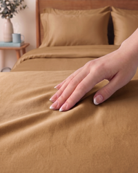 Hand touching golden sand percale sheet with wooden headboard and nightstand in the background