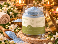 A glass jar of Jasmine Foot Soak filled with light blue crystals, shown with its lid leaning beside it on a cork coaster. The scene is styled with white flowers, a wooden spoon holding blue crystals, and warm candles in the background.