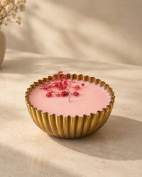 A pink scented candle in a fluted gold metal bowl, decorated with small pink dried flowers on top, set on a warm beige surface with soft shadows.