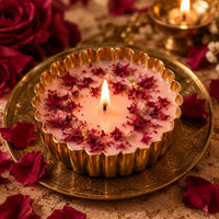 A lit pink soy candle in a fluted brass bowl, topped with dried red rose petals, resting on an engraved brass plate among red roses and petals with a brass diya glowing behind.